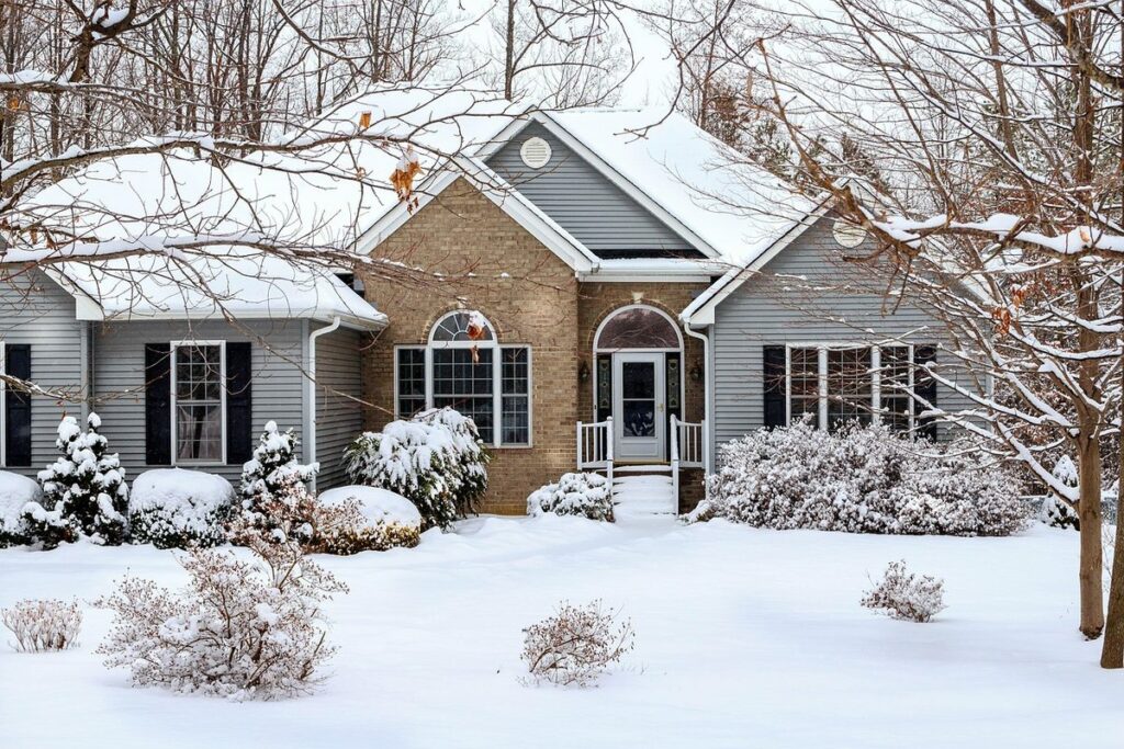 A gray and brick single-story house with a snow-covered roof, yard and shrubs